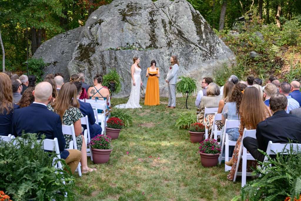 Outdoor wedding ceremony featuring a bride, groom and officiant in front of large bedrock of the Blue Ridge Mountains as the backdrop. Guests are seated theatre style in white wooden folding chairs with greenery arranged at the end of the rows, adding charm to the natural setting.